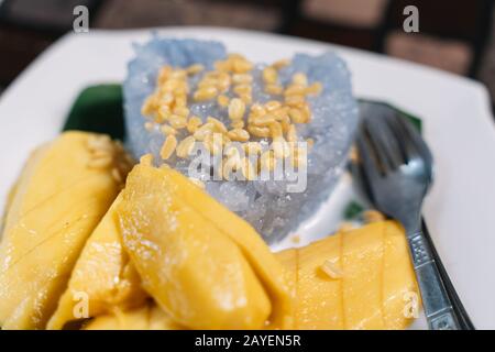 Traditionelles thailändisches Mango- und Reisgericht namens Mango Sticky Rice mit einer Gabel und einem Löffel auf einem Holztisch in Thailand. Gastronomie- und Reisekonzept Stockfoto