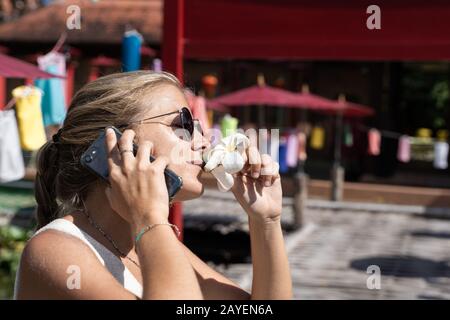 Blondes Mädchen mit Sonnenbrille, das eine weiße Lilie riecht, während sie auf ihrem Handy vor einem Gebäude in Thailand spricht. Reisekonzept Stockfoto