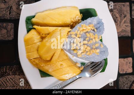Traditionelles thailändisches Mango- und Reisgericht namens Mango Sticky Rice mit einer Gabel und einem Löffel auf einem Holztisch in Thailand. Gastronomie- und Reisekonzept Stockfoto