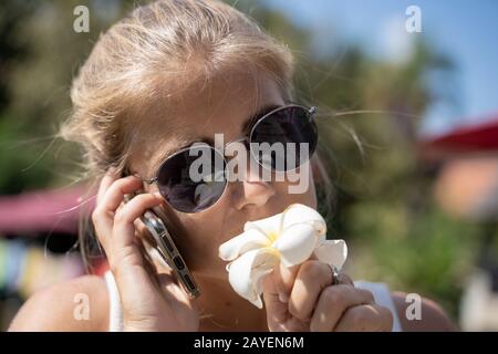 Blondes Mädchen mit Sonnenbrille, das eine weiße Lilie riecht, während sie auf ihrem Handy in Thailand spricht. Reisekonzept Stockfoto