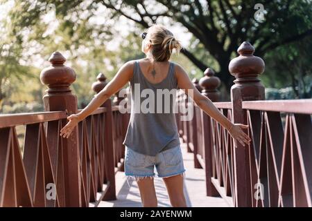 Junge blonde Frau von hinten auf einer Brücke in der Natur mit Tattoo auf dem Rücken mit ausgestreckten Armen, Freiheitsbegriff Stockfoto