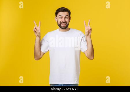 Junger gutaussehender Mann, der gestreiftes T-Shirt über isoliertem gelbem Hintergrund trägt und lächelt, als er auf die Kamera blickt, die die Finger zeigt, die V tun Stockfoto
