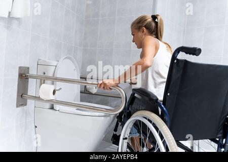 Blondes Mädchen, das sich auf der toilette an den Schienen hält und versucht, mit den Armen aus dem Rollstuhl aufzustehen, um zum Badezimmer zu gehen Stockfoto