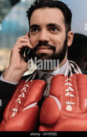 Vertikales Foto eines Geschäftsmannes in einem Anzug, der mit roten Boxhandschuhen am Hals an einer Glaswand hängt, auf seinem Handy spricht Stockfoto