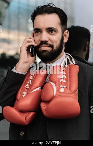 Vertikales Foto eines Geschäftsmannes in einem Anzug, der auf seinem Handy mit roten Boxhandschuhen spricht, die an seinem Hals hängen, der an eine Glaswand gelehnt ist Stockfoto