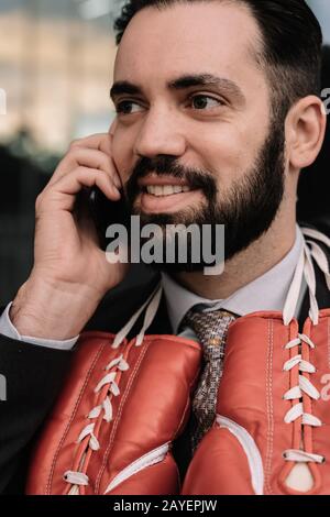 Vertikales Foto eines Geschäftsmannes in einem Anzug, der mit roten Boxhandschuhen am Hals an einer Glaswand hängt, auf seinem Handy spricht Stockfoto