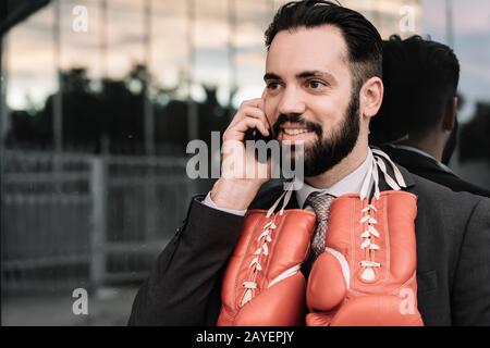 Geschäftsmann im Anzug mit roten Boxhandschuhen am Hals, der an eine Glaswand gelehnt ist, auf seinem Handy spricht Stockfoto