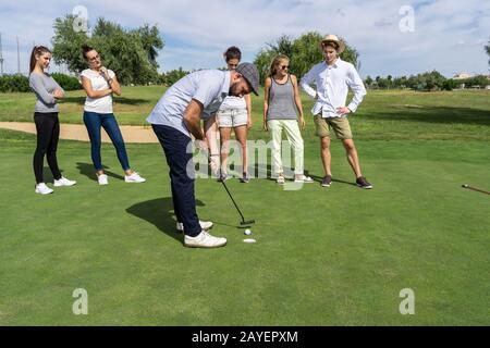 Junger Mann mit Bart und einem Golfschläger mit einem Golfclub vor einer Gruppe von Menschen, die ihn auf einem Golfplatz beobachten Stockfoto
