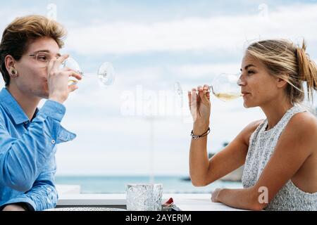 Junges Paar trinkt im Sommer Weißwein aus einem Glas, das auf einer Restaurantterrasse vor dem Meer sitzt Stockfoto