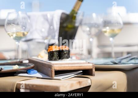 Sushi-Speisen mit Fischeiern werden auf einem kleinen Holzbrett in einem Tisch mit Weingläsern und einer Flasche Wein auf einer Terrasse eines Restaurants im Freien serviert. Stockfoto