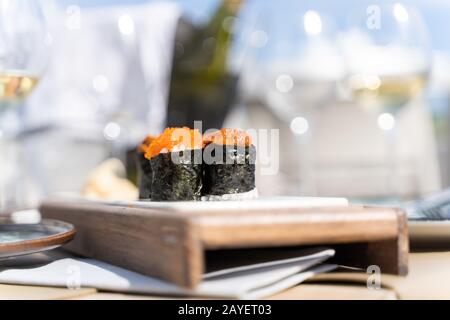 Sushi-Speisen mit Fischeiern werden auf einem kleinen Holzbrett in einem Tisch mit Weingläsern und einer Flasche Wein auf einer Terrasse eines Restaurants im Freien serviert. Stockfoto