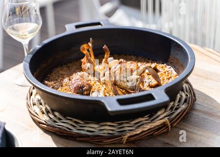 Reis mit Meeresfrüchten in einem Eisentopf auf einem Holztisch mit einem Glas Wein auf einer Terrasse eines Restaurants im Freien Stockfoto