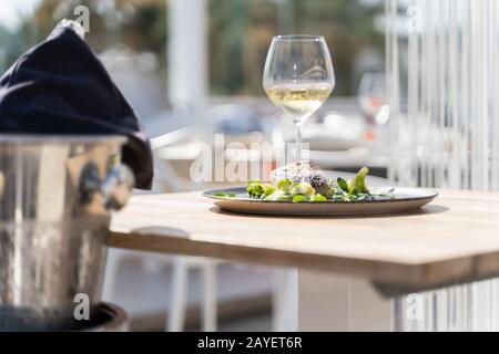 Fischgericht mit Gemüse wird in einem Holztisch mit Weingläsern und einer Flasche Wein auf einer Terrasse eines Restaurants im Freien serviert. Stockfoto