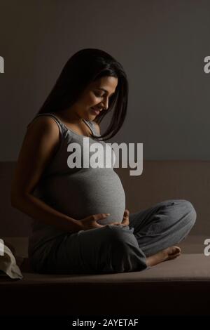 Schwangere Frau sitzt auf dem Bett und hält ihr Baby fest. Stockfoto
