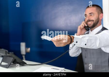 Stock-Foto eines Hotelempfängers, der am Telefon spricht und lächelt, während er seinen Arm mit einer Zimmerkarte in der Hand ausstreckt Stockfoto