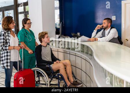 Stock-Foto eines jungen Jungen im Rollstuhl in Begleitung einer Krankenschwester und eines Mädchens mit einem Koffer an einer Krankenhausrezeption warten, während der Rezeptionist spricht Stockfoto