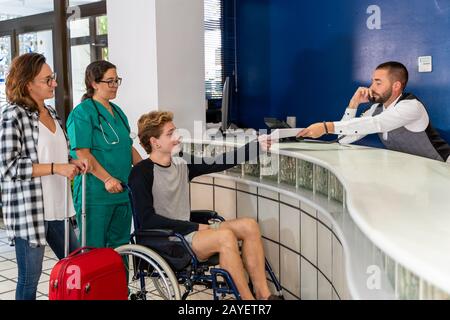 Stock-Foto eines jungen Jungen im Rollstuhl in Begleitung einer Krankenschwester und eines Mädchens mit einem Koffer an einer Krankenhausrezeption warten, während der Rezeptionist spricht Stockfoto