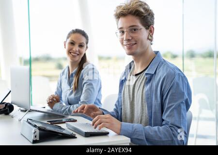 Junge Paare betrachten die Kamera und arbeiten an ihrem Computer in einer Feigheit Stockfoto