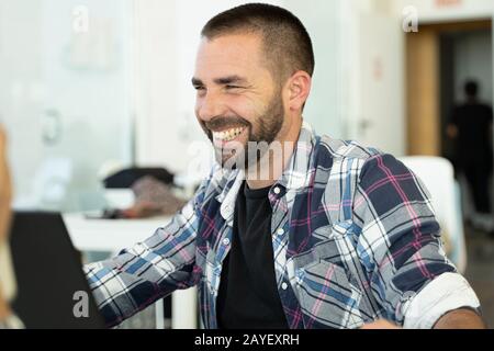 Junger Mann mit Bart und einem karierten T-Shirt, der an einem Tisch sitzt und mit anderen Leuten in einer Feigheit lacht Stockfoto