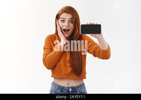 Amüsierte, aufgeregt hübsche Mädchen mit Rotkopf und orangefarbenem 3/4-Pullover, die den Kiefer aus Staunen seufzen und fallen lassen, während sie das Smartphone horizontal zeigen Stockfoto