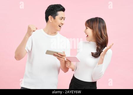 Zwei fröhliche Freunde stehen isoliert über rosafarbenem Hintergrund und spielen Spiele auf dem Handy. Stockfoto