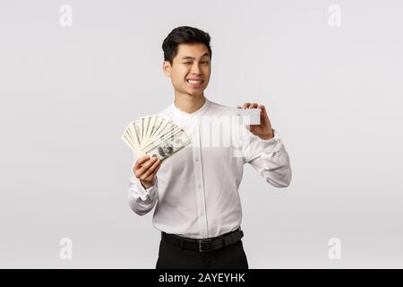 Entspannen Sie sich auf der Bank. Frech und entspannt, fröhlich erfolgreicher asiatischer Geschäftsmann winkt Kamera und lächelt, zeigt Geld und cr Stockfoto