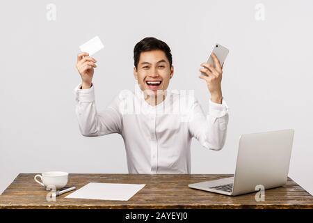 Mann feiert, Geste mit ja, hat viel gemacht, nach bestem Preis bestellt. Asiatischer Geschäftsmann hebt die Hände auf und hält Smartpho Stockfoto