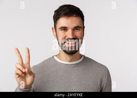 Nahestudio schoss fröhlichen, gutaussehenden bärtigen Mann in grauem Pullover, zeigt ein Friedenszeichen und lächelt sorglos, bleibt optimistisch und Opti Stockfoto