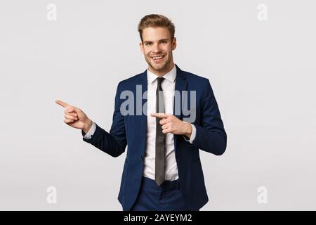 Stilvoller und selbstbewusster, gutaussehender blonder bärtiger Mann im klassischen blauen Anzug, nach links zeigend, der dem Geschäftspartner Platz zeigt, wo er Discs hat Stockfoto
