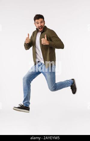 Hört sich gut an. Durchgehendes vertikales Studio schoss fröhlichen triumphierenden kaukasischen bärtigen Mann im Straßenoutfit und sprang aus Verwunderung Stockfoto