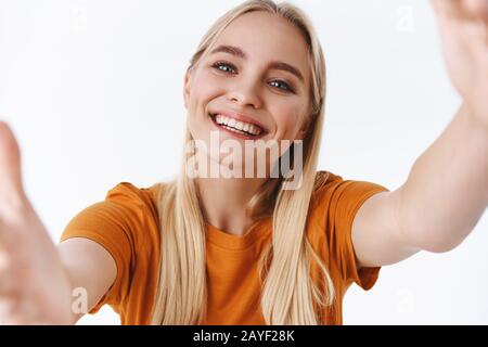 Nahaufnahme zärtliche, unbeschwerte blonde Frau mit Tätowierungen, orangefarbenes T-Shirt, halten Sie die Kamera mit beiden Händen seitlich, während Sie selfie nehmen und Videokonferenzen nutzen Stockfoto