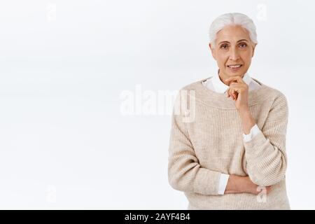 Neugierige, gut aussehende Seniorin, alte Frau in modischem eleganten Outfit, Pullover über Bluse, berührendes Kinn und Look Kamera fasziniert, lächelnd Stockfoto