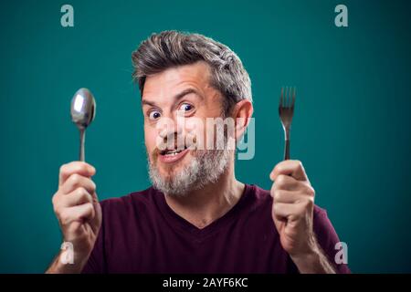 Ein Porträt eines bärtigen hungrigen Mannes, der Löffel und Gabel in den Händen hält. Menschen- und Lebensmittelkonzept Stockfoto