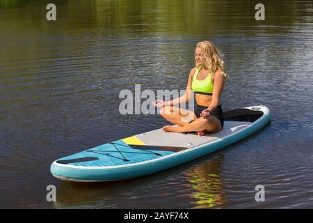 Junge blonde Frau meditiert auf SUP im Wasser Stockfoto