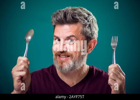 Ein Porträt eines bärtigen hungrigen Mannes, der Löffel und Gabel in den Händen hält. Menschen- und Lebensmittelkonzept Stockfoto