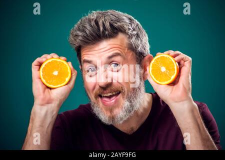 Ein Porträt eines bärtigen, lustigen Mannes in rotem T-Shirt, das die geschnittenen Orangen hinter dem Gesicht hält. Menschen, Emotionen und Essenskonzept Stockfoto