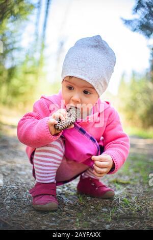 Adliges Baby, das sich auf dem Waldweg in der Natur duckt. Porträt eines 1-jährigen Mädchens im Wald mit Kiefernkegel im Monat. Stockfoto