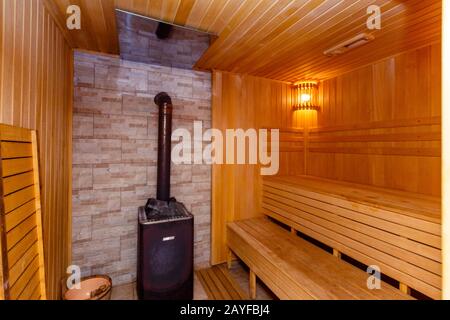 Die Sauna ist von Holz gesäumt. Schwitzraum. Herd. Heizung mit Steinen für Dampf. Regale zum Sitzen und Dampfgaren. Beige Fliesen an den Wänden Stockfoto