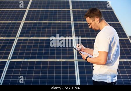 Seitenansicht eines jungen erfolgreichen Mannes, der an einer PV-Solaranlage steht und in seinem Ordner über einen Geschäftsplan nachdenkt und Notizen macht, Seitenansicht, alternatives Energiekonzept Stockfoto