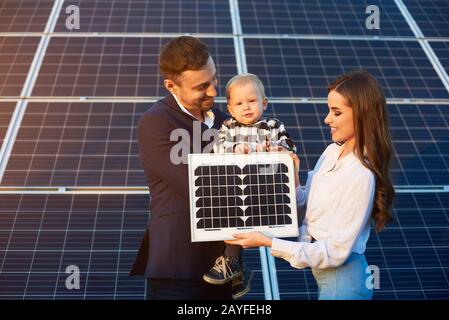 Der junge Vater hält seinen Sohn, die Mutter hält kleines pv-Modul, großes Solarmodul im Hintergrund, Konzept moderner Technologien Stockfoto