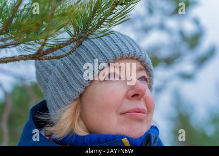 Frauen tragen einen blauen Strickhut. Fröhlich reife Frau lächelnd. Außenaufnahmen bei kaltem Wetter. Nahaufnahme der Druckseite. Kiefernbaum Hintergrund Stockfoto