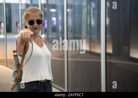 Blondes Mädchen mit Sonnenbrille, einem Geflecht und einem weißen Tanktop stehend und vor einem modernen Gebäude mit Glasfassade in Singapur zeigend Stockfoto