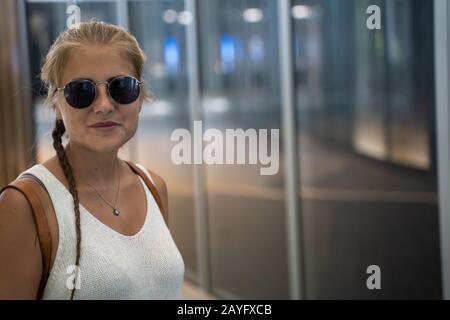 Blondes Mädchen mit Sonnenbrille, einem Geflecht und einem weißen Tanktop, das vor einem modernen Gebäude mit Glasfassade in Singapur steht Stockfoto