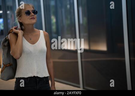 Blondes Mädchen mit Sonnenbrille, einem Geflecht und einem weißen Tanktop, das eine Tasche mit der Hand auf dem Rücken hält, das vor einem modernen Gebäude steht und einen sieht Stockfoto