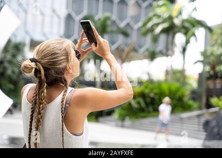 Blondes Mädchen mit Sonnenbrille und weißem Tanktop und einem Geflecht, das mit ihrem Handy ein Bild in einige moderne Gebäude in Singapur macht. Reise Konz Stockfoto