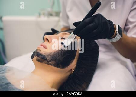 Der Kosmetologe in pinkfarbenen Handschuhen mit Pinsel trägt eine Kohlemaske zum Schälen auf das Gesicht eines jungen Mädchens in einem Kosmetologie-Raum auf. Das Konzept des Kosms Stockfoto