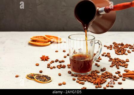 Frisch gebrühter Espressokaffee wird aus einer Cezve in eine transparente Glasbecher vor grauem Betongrund mit Kaffeebohnen gegossen, getrocknet Stockfoto