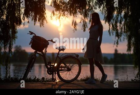 Silhouette einer Frau im Kleid und Retro-Fahrrad mit einem Korb unter Baumzweigen vor dem Hintergrund eines Sees und der Sonne bei Sonnenuntergang. Magische Sonnenuntergangslandschaft Stockfoto