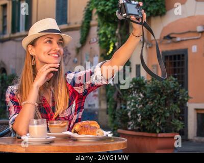 Junge blonde Frau mit blauen Augen, die beim Frühstück Selfies in der typischen italienischen Bar außerhalb des historischen Viertels Trastevere in Rom, Italien, nimmt Stockfoto