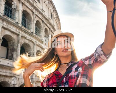 Junge blonde Frau mit blauen Augen und langen Haaren, wobei selfie mit einem Vintage Kamera am Kolosseum, Rom. Großen Hut, rot kariert Hemd. Stockfoto